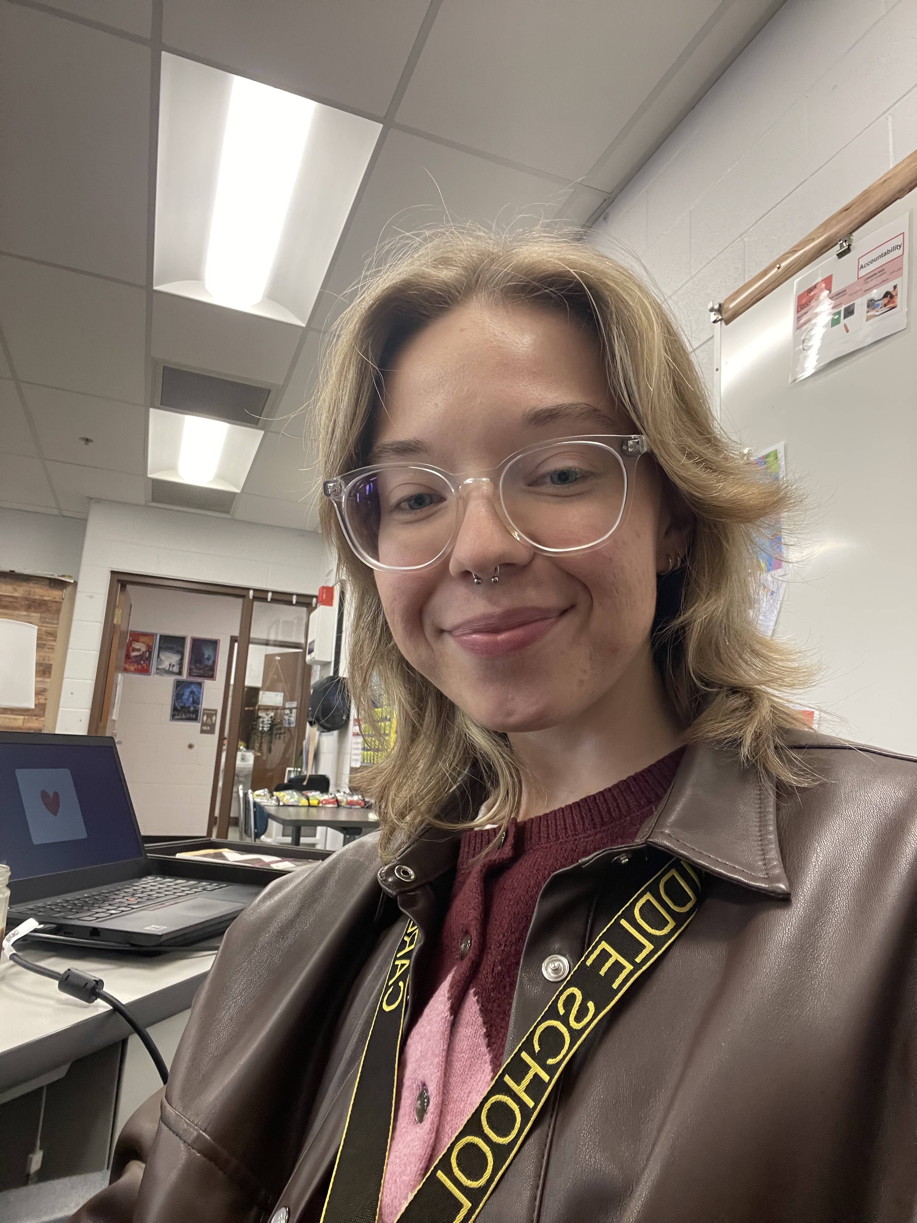An image of Aleks smiling for the camera. They have blonde hair, glasses, and are wearing a brown leather jacket.