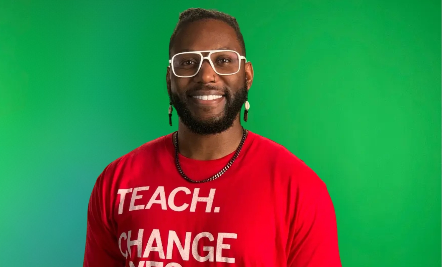 An image of Corey in front of a green screen smiling. He is wearing a red t-shirt that says "Teach. Change Lives."