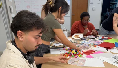 Students gather around a table cutting paper and assembling collages.