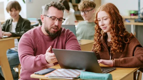 A secondary teacher discusses work on a laptop with a student.