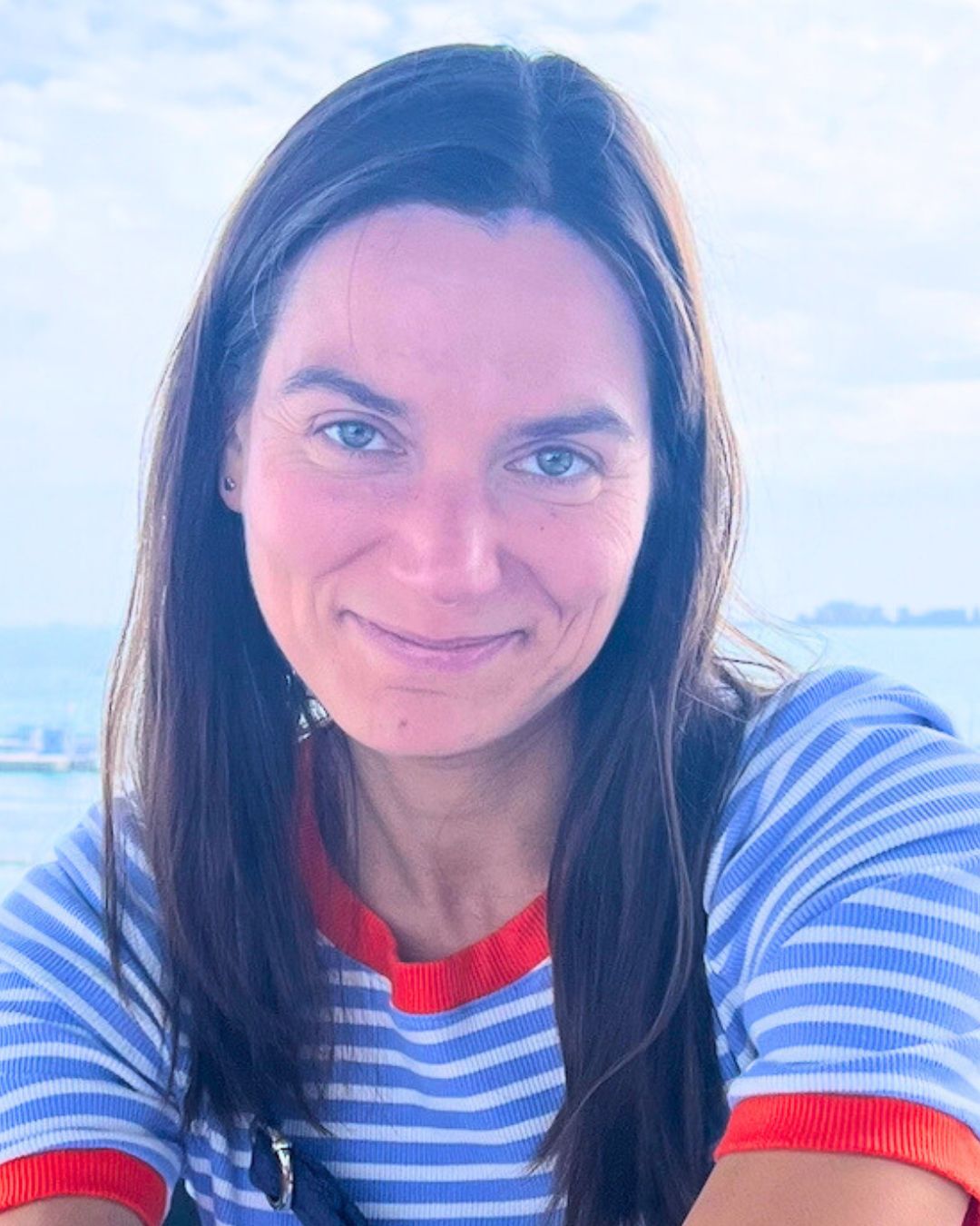 An image of Olga Vaskova smiling for the camera. She has long brown hair and is sitting in front of a body of water.