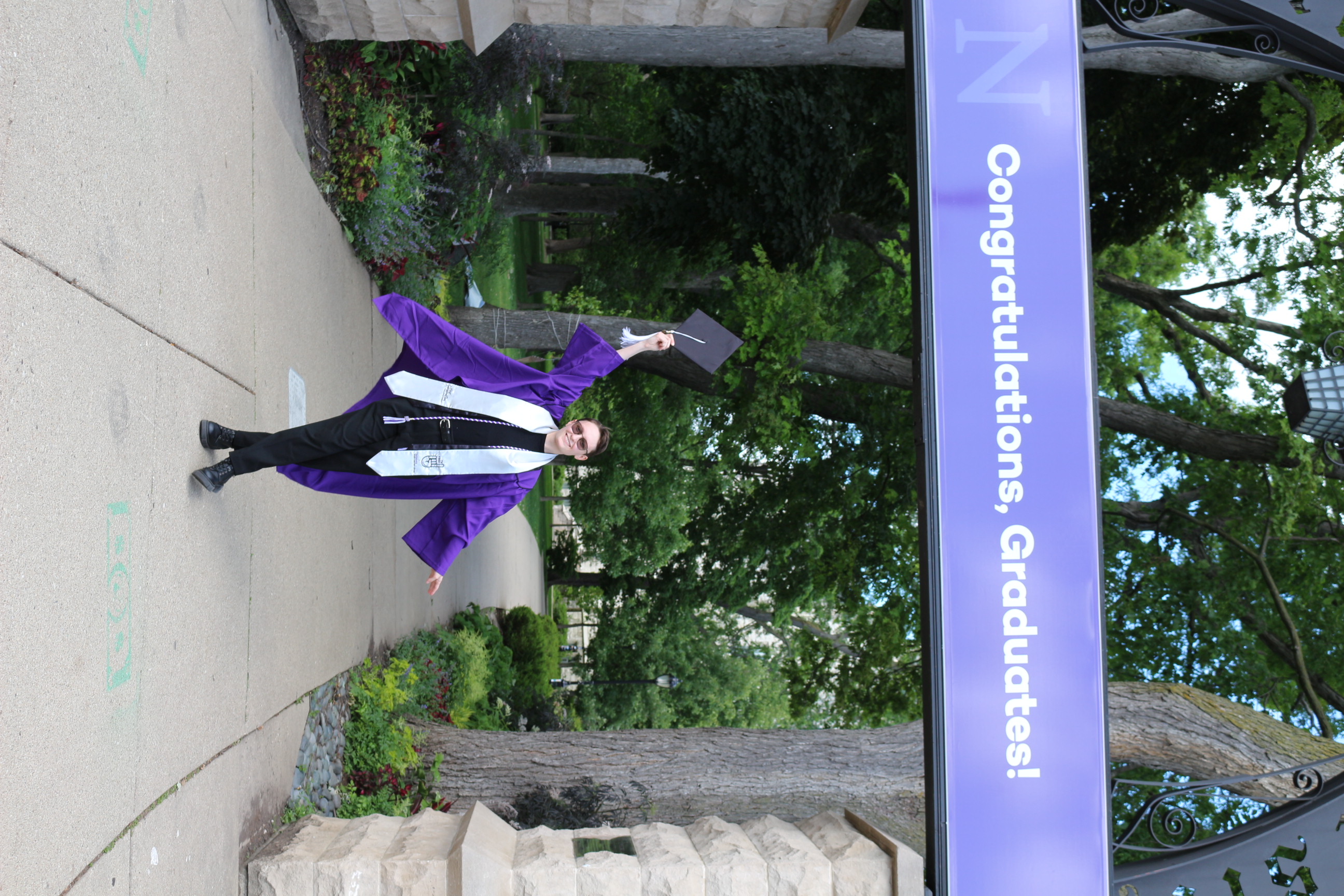Sylvia proudly standing under the arch in their cap and gown, upon which there is a sign that says "Congratulations, Graduates!"