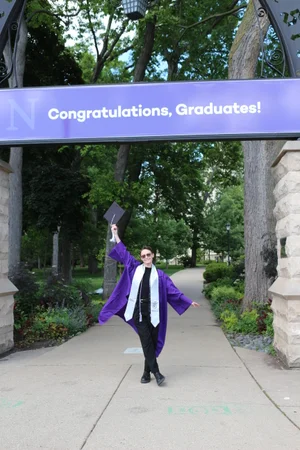 Sylvia proudly standing under the arch in their cap and gown, upon which there is a sign that says "Congratulations, Graduates!"