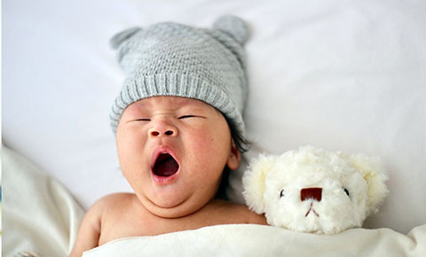 baby yawning in bed next to teddy bear